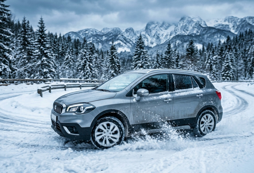 Srebrni Suzuki SX4 S-Cross crossover na dinamičnoj fotografiji u vožnji na snijegom prekrivenoj, zavojitoj planinskoj cesti. Automobil skreće ulijevo, podižući snijeg svojim kotačima. U pozadini su visoke, snijegom prekrivene planine i gusta crnogorična šuma pod tmurnim, oblačnim nebom.