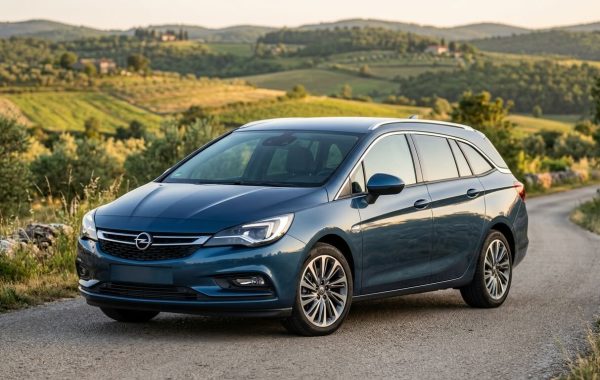 lavi karavan marke Opel Astra parkiran na zavojitoj asfaltiranoj cesti u ruralnom, brdovitom krajoliku sa suhozidima i stablima maslina u vrijeme zalaska sunca.
