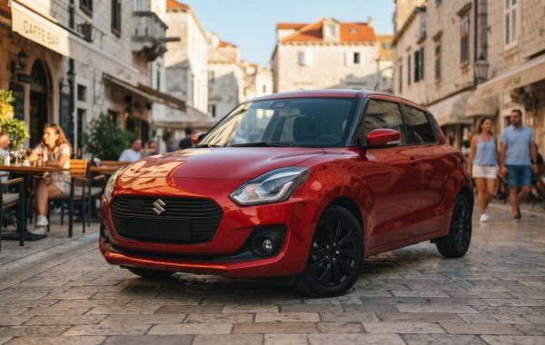 Fotografija prikazuje moderni crveni hatchback Suzuki Swift, parkiran na popločenoj ulici u povijesnom mediteranskom gradu. Automobil je usmjeren prema naprijed i blago udesno, a njegovi prepoznatljivi prednji farovi i crni aluminijski naplatci jasno su vidljivi. U pozadini, na kamenoj fasadi zgrade, nalazi se natpis "CAFFE BAR", koji otkriva da se radi o kafiću s terasom. Nekoliko ljudi sjedi za stolovima na terasi s lijeve strane, dok par šeće ulicom u pozadini s desne strane.